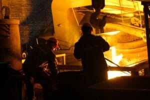 Two workers operate in a steel factory, managing molten metal in an industrial furnace.