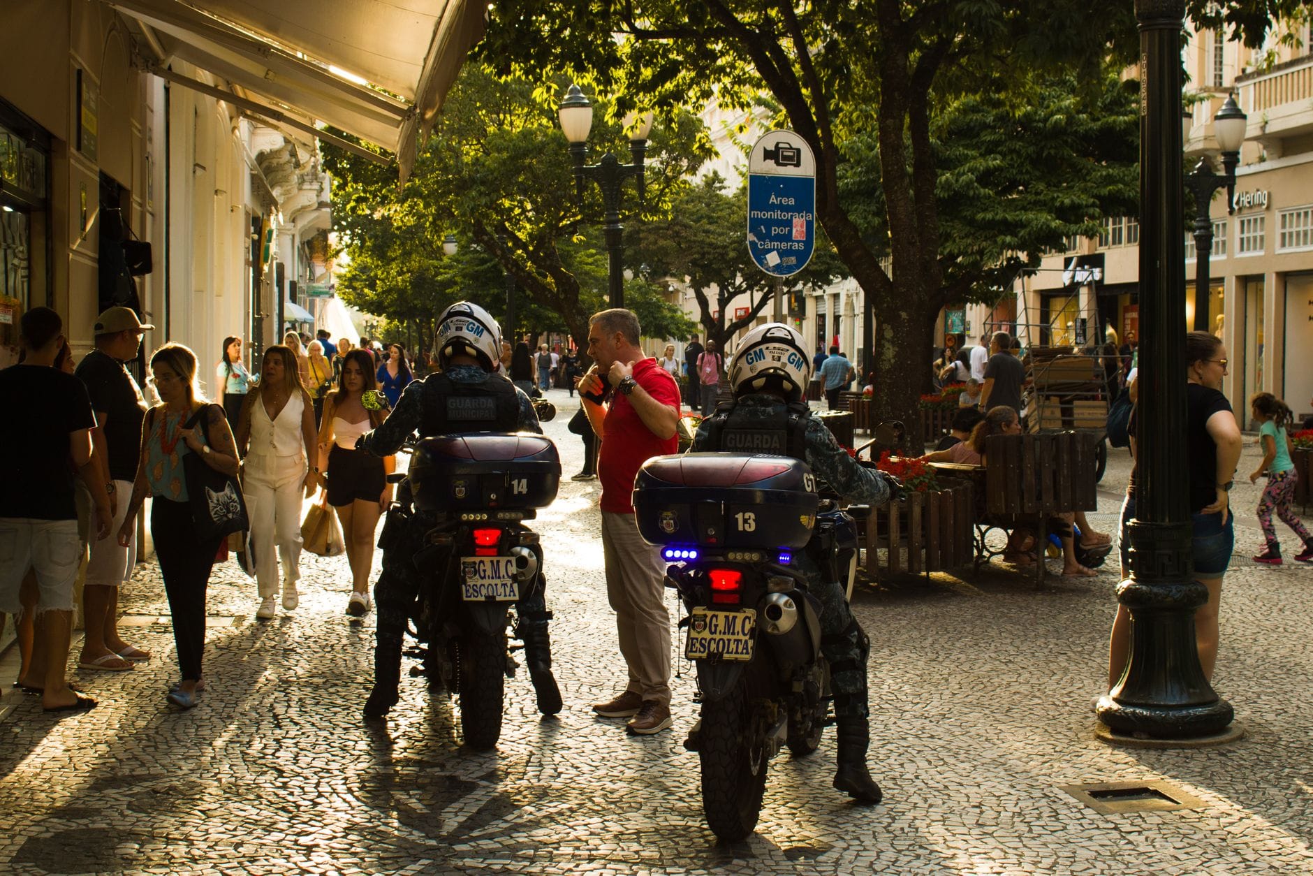 Municipal guards on motorcycles patrol a lively street in Brazil with bustling pedestrian activity.