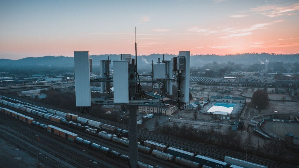 Drone view of contemporary cell tower located near railway tracks with cargo containers and wagons against sunset sky