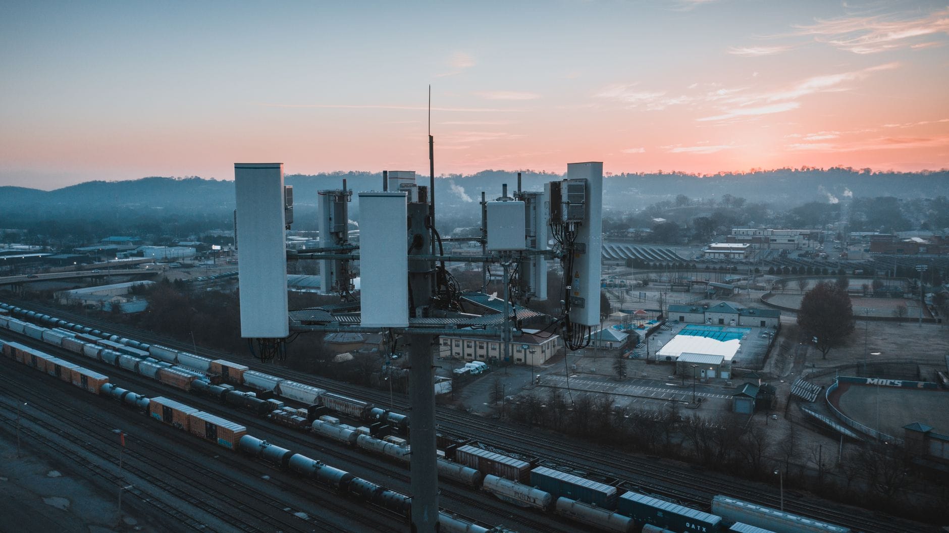 Drone view of contemporary cell tower located near railway tracks with cargo containers and wagons against sunset sky